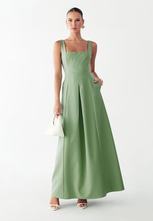 Vrouw in een mouwloze, groene jurk tot op de grond, met een witte handtas, witte hakken en gouden oorringen.