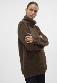 Pull à col roulé oversize marron en texture moelleuse avec des poignets côtelés, présentant de subtils éclats multicolores. Porté avec un jean noir.