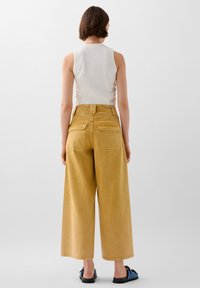 Pantaloni gialli a gamba larga con vita alta, due tasche posteriori e una finitura testurizzata, abbinati a una canotta bianca aderente e scarpe blu.