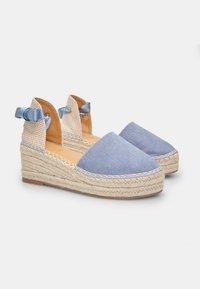 Modré plátenné espadrilky so splietanými juty podrážkami, ktoré majú textúrovanú béžovú látku na chrbte a svetlomodré stuhy ako ozdobu.