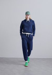Marineblauwe hoodie met witte trekkoorden en een zak aan de voorkant, gecombineerd met bijpassende joggers. Een groene pet en felle groene schoenen maken de outfit compleet.