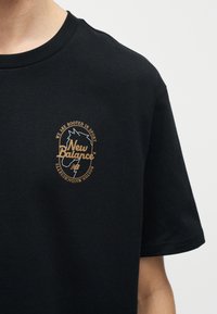 Μαύρο T-shirt με κοντά μανίκια, με μικρό μπεζ λογότυπο New Balance και γραφικό κεφάλι αλόγου στο αριστερό στήθος.