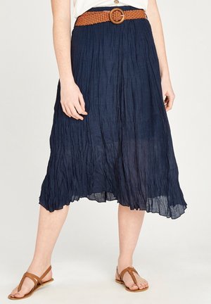Vrouw draagt een geplooide navy midi-rok met een gevlochten bruine riem en bruine gevlochten platte sandalen, staand met de benen licht uit elkaar.