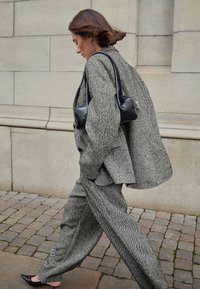 Grey textured oversized suit with a tailored fit, paired with a small black handbag and pointed black shoes, walking on cobblestone.