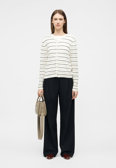 Γυναίκα που στέκεται, φοράει λευκή ριγέ ζακέτα, μαύρο παντελόνι με φαρδιά πόδια, καφέ παπούτσια και κρατάει μια μπεζ τσάντα στο πλάι της.