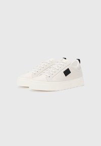 Baskets en toile blanches avec un design texturé, dotées de lacets plats, d'une pièce noire en accent et d'une épaisse semelle blanche. Style minimaliste.