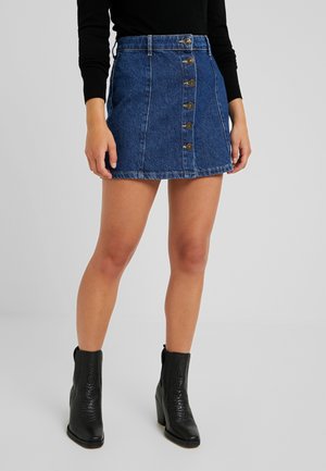 Kvinde iført en blå denim mininederdel med knapper foran, sort langærmet top og sorte ankelstøvler med chunky hæle.