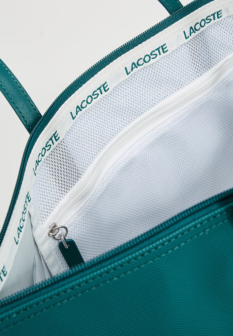 Interior de bolso de tote texturizado verde con un forro blanco que incluye un bolsillo de malla con cremallera y el branding repetido "LACOSTE" en el borde.
