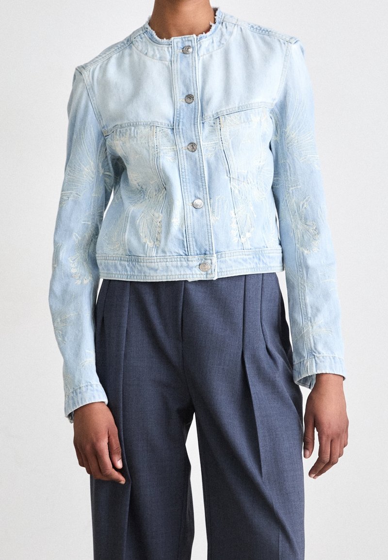 Personne portant une veste en jean bleu clair avec broderie et boutons métalliques, assortie à un pantalon plissé taille haute gris foncé.