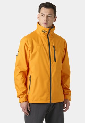 Man die een feloranje jas met rits en hoge kraag draagt, met zwarte ritssluitingen, staand tegen een effen achtergrond.