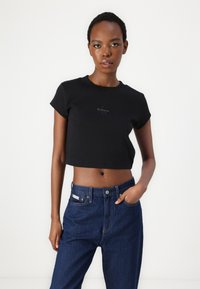 Melnā īsā t-kreklā ar ribotu tekstūru un īsām piedurknēm, kam priekšpusē ir neliels "Calvin Klein" logo, apvienots ar ziliem džinsiem.
