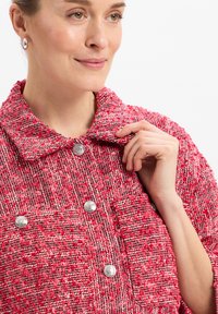 Rote strukturierte Jacke mit großem Kragen und silbernen Knöpfen. Der Stoff hat ein gewebtes Muster mit verschiedenen Rot- und Weißtönen.