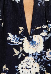Robe fleurie avec un décolleté en V profond ; fond bleu marine foncé avec des fleurs bleu clair et crème ; tissu texturé avec un détail plissé.