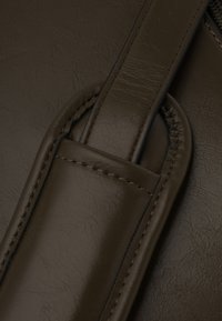 Yksityiskohta ruskeasta nahkalaukusta, jossa on sileä pinta, ommellut korostukset ja pyöreä kahvakiinnitys, jossa on hienovarainen hieno kuvio.