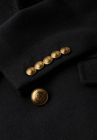 Zwarte wollen blazer met vijf gouden knopen met reliëfontwerpen en een grotere gouden knop met gegraveerde tekst aan de voorkant.