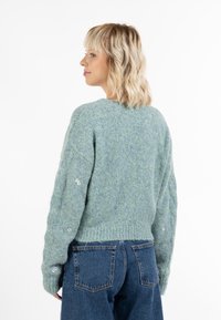 Ljusgrön stickad tröja med en texturerad finish, ribbad nederkant och dekorerade broderade blommor på ärmarna. Bärs med blå denimjeans.