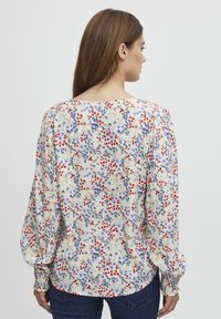 Blusa a stampa floreale con maniche a sbuffo lunghe, caratterizzata da uno sfondo crema e fiori colorati in rosso, blu e verde. Tessuto leggero.