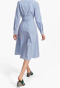 Sininen ja valkoinen raidallinen paita-mekko, jossa on istuva vyötärö ja levenevä hame. Pitkät hihat napillisten hihansuiden kanssa. Vihreät ja mustat korokepohjaiset sandaalit.