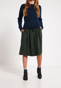 Suéter de punto azul oscuro combinado con una falda de ante verde y fluida, con cintura elástica y cordón, llevada con botas negras por los tobillos.