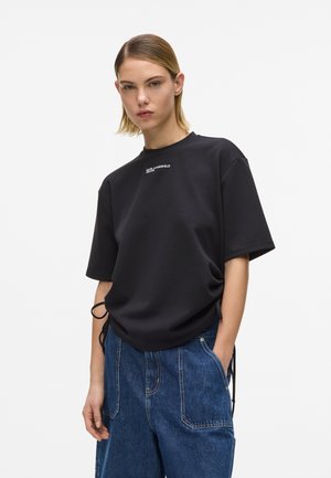 Młoda osoba ubrana w czarny, oversize'owy t-shirt z białym logo oraz luźne niebieskie dżinsowe spodnie, stojąca z jedną ręką w kieszeni.