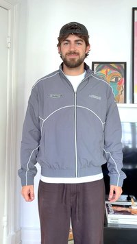 Mees, kes kannab halli lukuga jope valgete äärtega, musta pesapalli nokamütsi ja tumedaid pükse, seisab siseruumis abstraktse kunsti ja televiisori ees.