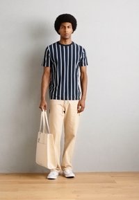 Dryžuota tamsiai mėlyna ir balta trumpomis rankovėmis marškiniai su šviesiai beige kelnėmis. Modelis laiko šviesiai beige rankinę ir dėvi baltus batus. Lygus audinys.