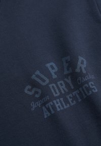 Donkerblauwe sweatshirt met gestructureerde stof, ronde logo in lichtblauwe tekst: "SUPER DRY JAPAN OSAKA ATHLETICS" met een strak ontwerp.
