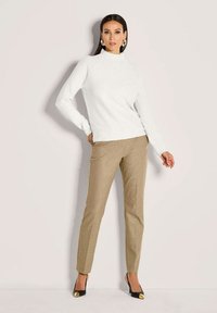 Frau trägt einen weißen Rollkragenpullover, beige schmal geschnittene Hosen, schwarze Schuhe mit goldenen Akzenten und goldene Creolen, steht vor einer schlichten Wand.