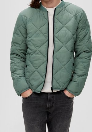 Homme portant une veste matelassée zippée vert clair sur une chemise blanche, les mains dans les poches, jean noir visible de la taille vers le bas.