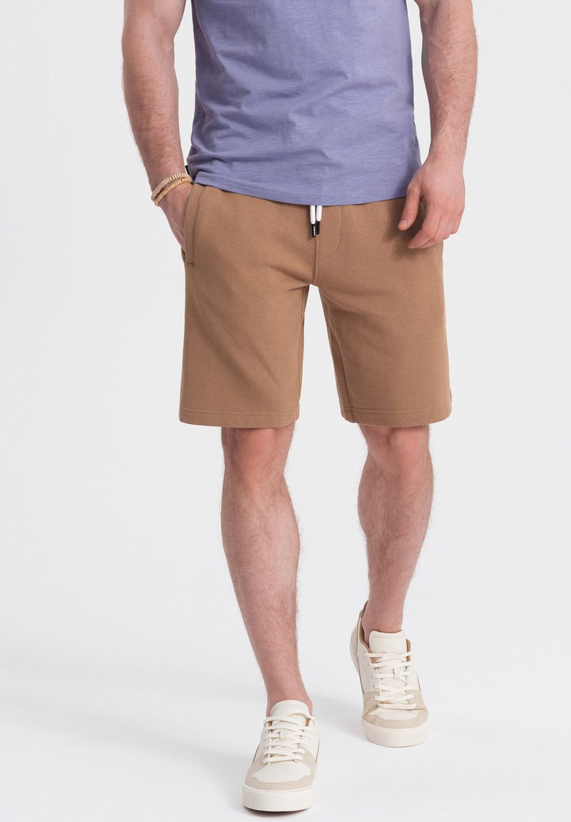 Shorts en coton beiges avec une taille élastique et un cordon de serrage, dotés de poches latérales, associés à des baskets claires et à une chemise lavande.