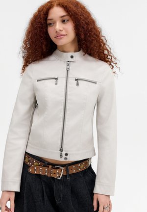 Jonge vrouw met krullend rood haar die een lichtgrijs jack met rits en een donkere broek draagt met een bruine gespenriem.