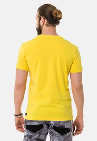Gelbes Kurzarm-T-Shirt aus Baumwolle mit Rundhals. Getragen mit grauen gemusterten Hosen. Einfach, einfarbig mit glatter Textur.