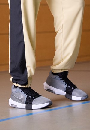 Sneaker atletici grigi e neri realizzati in mesh e materiali tessili, con suola bianca e chiusura con lacci, indossati con pantaloni gialli e neri.
