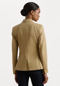 Blazer beige sur mesure avec épaules structurées, fente au dos et accents de boutons dorés aux poignets. Tissu texturé, design ajusté.