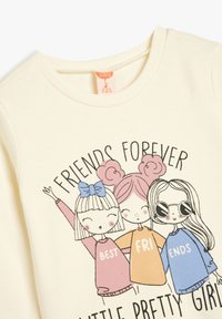 Sweatshirt en coton crème présentant trois filles de dessin animé avec des tenues colorées. Le texte indique "AMIES POUR TOUJOURS" au-dessus de leurs sourires animés.