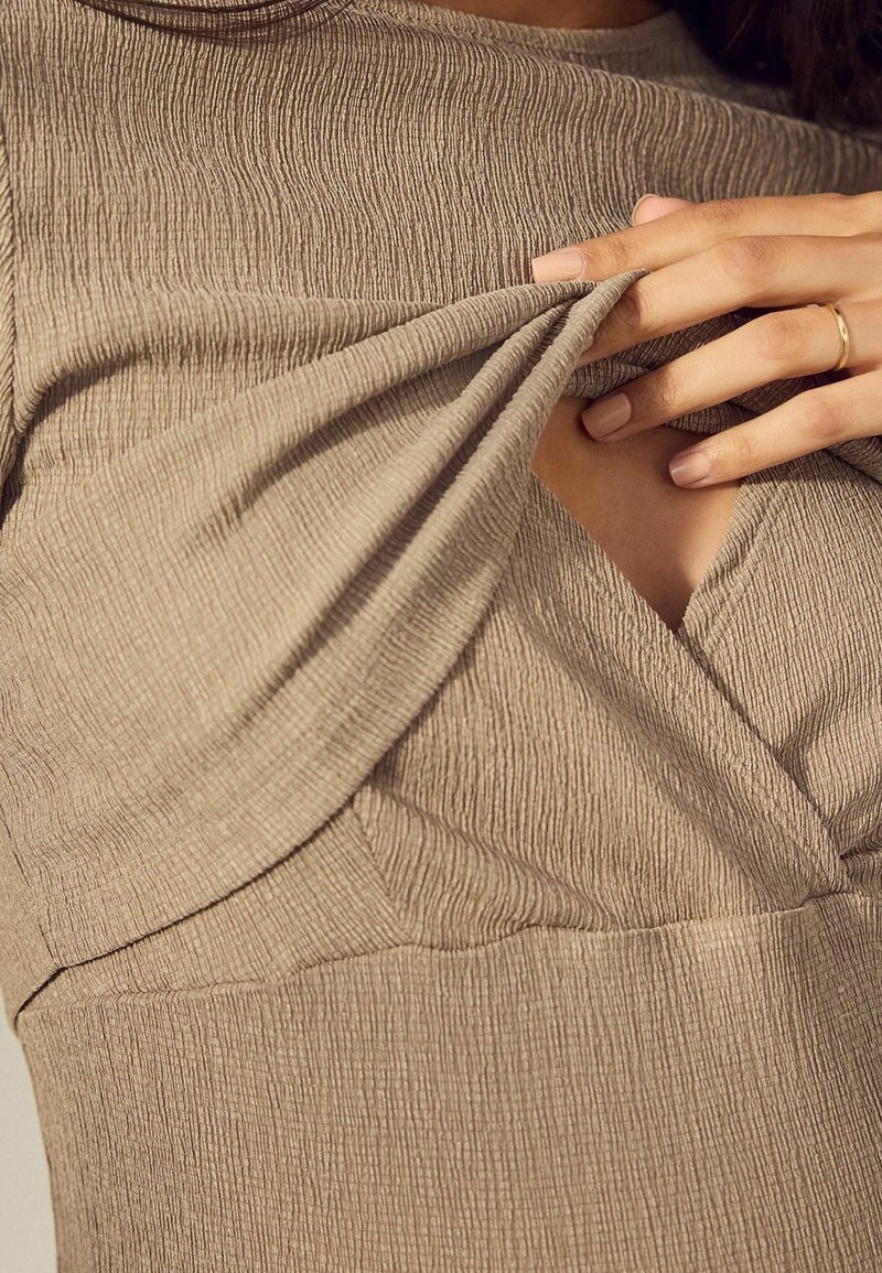 Robe marron texturée avec un corsage ajusté et un détail croisé sur la poitrine. Le tissu présente un motif côtelé et une légère élasticité.