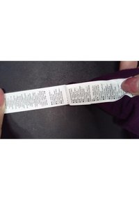 Tvättvårdsetikett av vitt tyg, med svart text. Innehåller en lista över material, tvättanvisningar och plaggdetaljer, med en vikning i kanten.