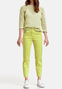 Světle žlutý pletený svetr s tříčtvrtečními rukávy a kulatým výstřihem, doplněný jasně zelenými zúženými kalhotami a bílými teniskami.