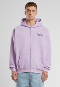 Ljubičasta zip-up jakna s prednjim džepom, rebrastim manžetama i pojasom. Sadrži "KANI Music" logo u crnoj boji na lijevoj strani prsnog područja.