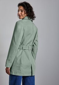 Jasnozielony trench z paskiem w talii, tylnym rozcięciem i strukturalnym kołnierzem, połączony z niebieskimi dżinsami o szerokich nogawkach. Gładka tekstura materiału.