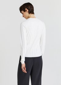 Weißes geripptes Langarmshirt mit figurbetontem Schnitt, Rundhalsausschnitt und strukturiertem Stoff. Getragen mit dunklen Hosen, seitliche Ansicht.