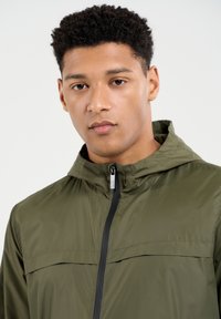 Alyvuogių žalios spalvos lengvas švarkelis su užtrauktuku, gobtuvu ir juodais akcentais; glotni tekstūra ir laisvas kirpimas, su horizontaliu krūtinės kišenė.