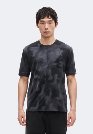 Junger Mann, der nach vorne schaut, trägt ein schwarz-dunkelgraues T-Shirt mit Camouflage-Muster und schwarze Hose vor einfachem Hintergrund.