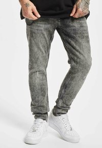 Bărbat îmbrăcat cu blugi skinny negri decolorati, cu ușoare deteriorări și adidași albi Nike, cu mâinile tatuate odihnindu-se lângă talie.