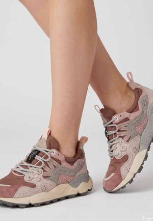 Gambe con sneakers chunky rosa e grigie con dettagli decorati e linguette, in piedi su un pavimento grigio chiaro contro uno sfondo semplice.
