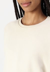 Sudadera color crema con cuello redondo acanalado y textura suave, con un corte relajado y sin gráficos ni adornos visibles.