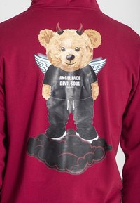 Bordo dukserica s grafikom medvjeda u crnoj košulji i hlačama, s krilima i rogovima, koji stoji na oblaku s tekstom ispod.