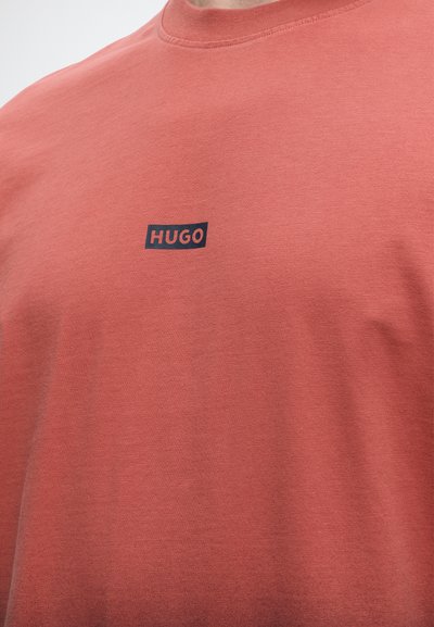 Детайл на човек, носещ риза в цвят ръжда с малко черно и червено лого "HUGO" на гърдите.