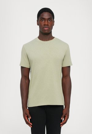 Tânăr stând cu fața spre înainte, îmbrăcat cu un tricou cu mânecă scurtă de culoare verde deschis și pantaloni negri, pe un fundal alb simplu.