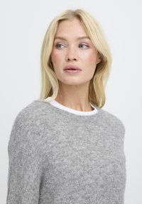 Pulover gri realizat dintr-un material moale, texturat, cu guler rotund și o cămașă albă stratificată dedesubt. Designul este de tip lejer.